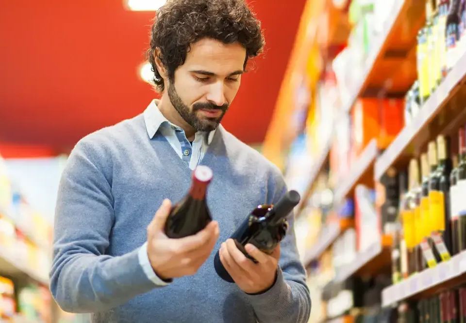 Man in a light blue sweater compares two wine bottles in a store aisle, considering the psychology of pricing.