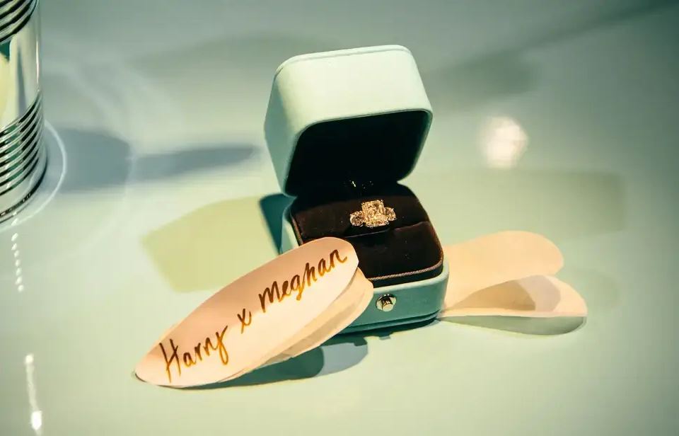 Diamond engagement ring in an open Tiffany blue box next to a white card with 'Harry x Meghan' written in gold, showcasing pricing psychology.