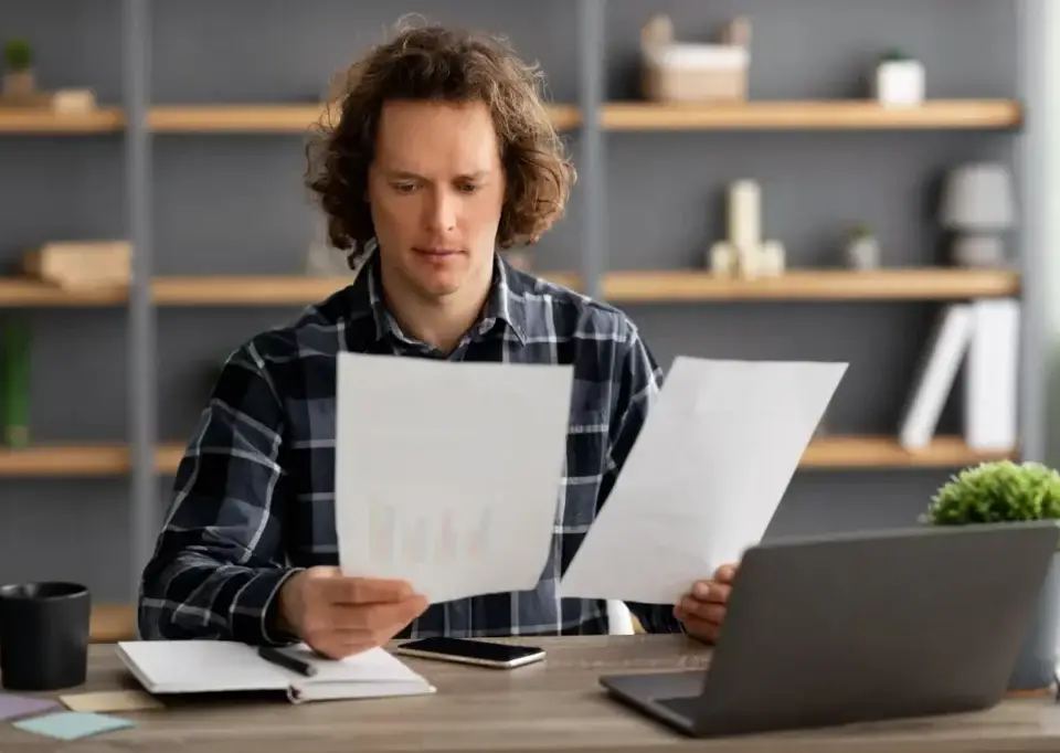 Man with curly hair reviewing documents at a desk, laptop and coffee nearby, considering fractional leadership.