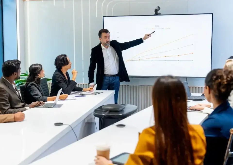 Manager pointing at a sales graph on a large monitor, explaining what CPA in digital marketing means to his diverse team.